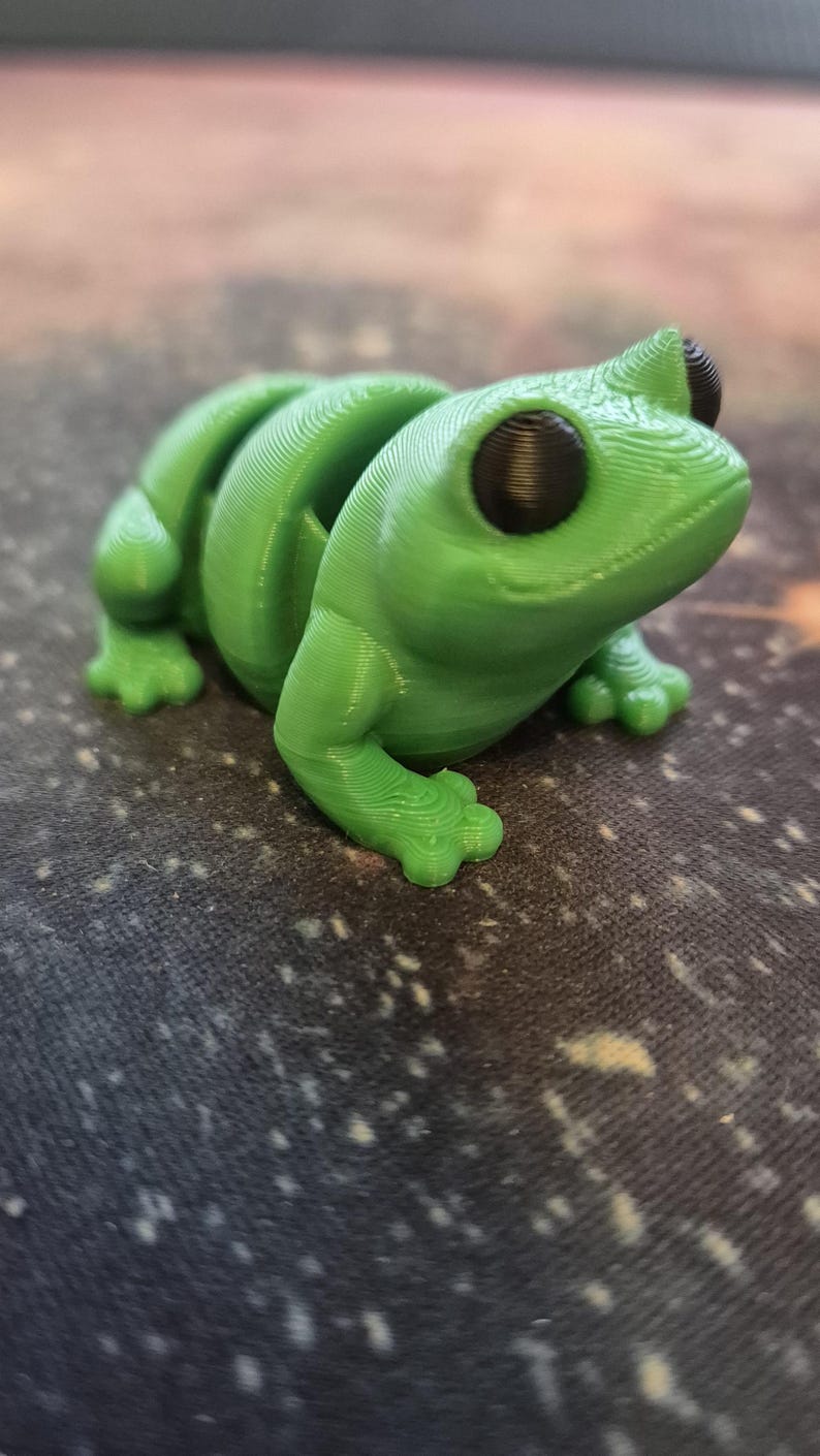 May include: A 3D printed green frog figurine with black eyes. The frog is sitting on a dark fabric surface with a speckled pattern.