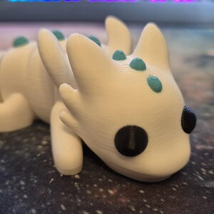 May include: A white 3D printed dragon figurine with teal accents and black eyes. The dragon is lying on its belly with its head turned to the side.