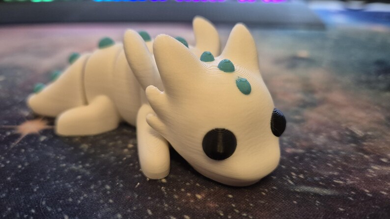 May include: A white 3D printed dragon figurine with teal accents and black eyes. The dragon is lying on its belly with its head turned to the side.