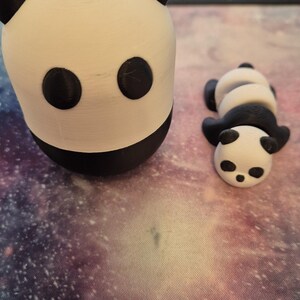 May include: Two 3D-printed panda figurines. One is upright, the other is lying down. Both are white with black accents for the ears, eyes, and body. The figurines are on a galaxy-themed surface.