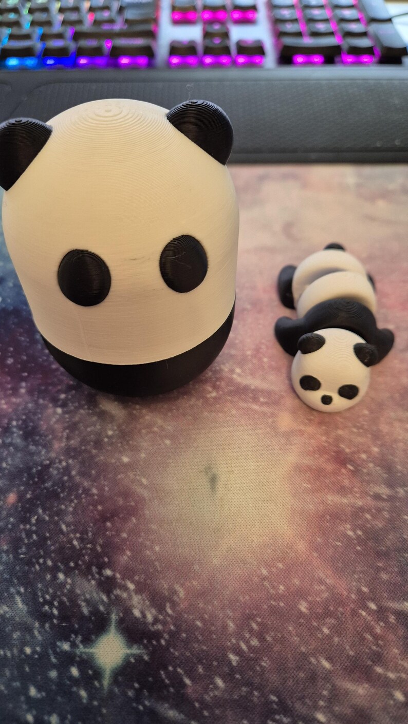May include: Two 3D-printed panda figurines. One is upright, the other is lying down. Both are white with black accents for the ears, eyes, and body. The figurines are on a galaxy-themed surface.