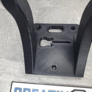 May include: Black plastic wall mount with a guitar-shaped cutout. The mount has two curved arms and a flat base.