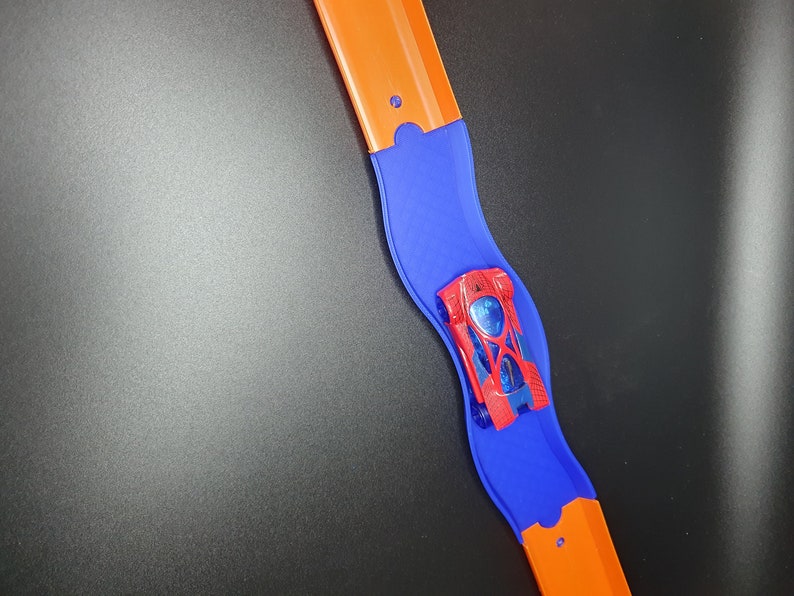 May include: A red toy car with blue accents is on a blue and orange track. The track is curved and the car is going around the curve.