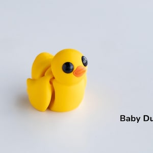 May include: A small yellow rubber duck with black eyes and an orange beak. The duck is sitting on a white surface. Baby Duck.