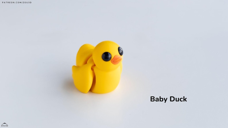 May include: A small yellow rubber duck with black eyes and an orange beak. The duck is sitting on a white surface. Baby Duck.