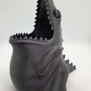 May include: A black dinosaur-shaped planter with a textured finish. The planter has an open mouth and a small eye detail.