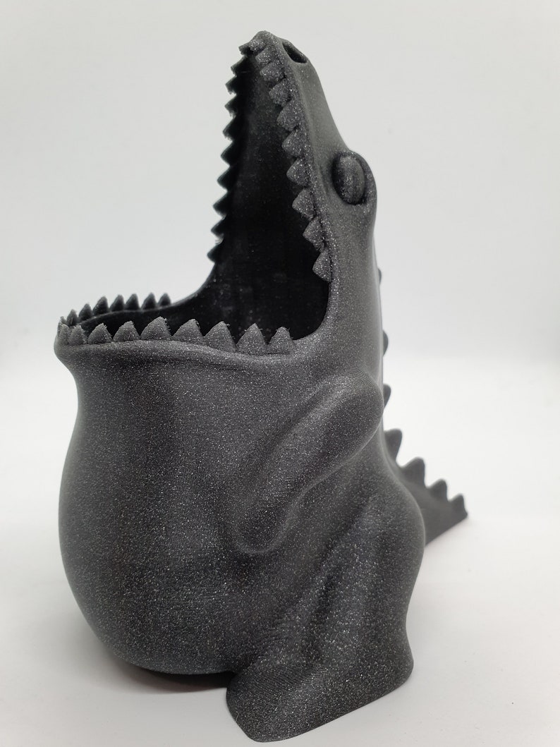 May include: A black dinosaur-shaped planter with a textured finish. The planter has an open mouth and a small eye detail.