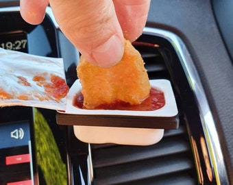 Puede incluir: Un recipiente de plástico blanco con salsa roja y una nugget de pollo que se está sumergiendo en él. El recipiente está unido a la ventilación de aire de un coche.
