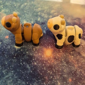 May include: Two 3D-printed articulated animal figures. One is brown and resembles a horse, the other is white with black spots, resembling a cow. Both have black eyes and feet. The figures are on a galaxy-themed surface.