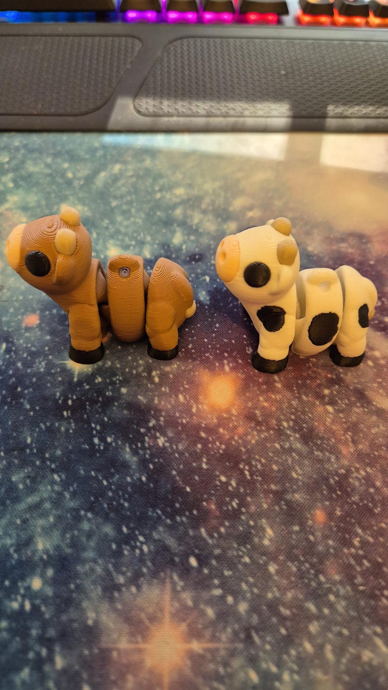 May include: Two 3D-printed articulated animal figures. One is brown and resembles a horse, the other is white with black spots, resembling a cow. Both have black eyes and feet. The figures are on a galaxy-themed surface.