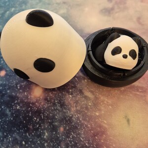 May include: A white panda-shaped container with three black spots and a separate black container holding a panda figurine. The panda figurine has black ears and eyes. The items are on a galaxy-themed surface.