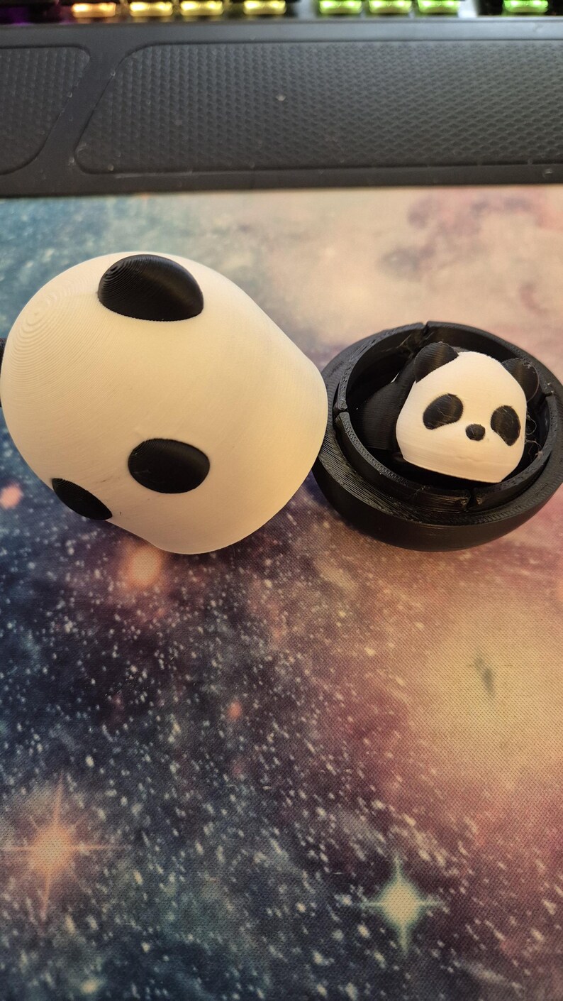May include: A white panda-shaped container with three black spots and a separate black container holding a panda figurine. The panda figurine has black ears and eyes. The items are on a galaxy-themed surface.