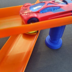 Puede incluir: Un coche de juguete rojo con ventanas tintadas azules est&aacute; en una pista de pl&aacute;stico naranja. La pista est&aacute; conectada a una base de pl&aacute;stico azul.