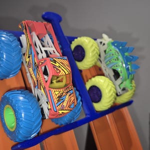 May include: Three toy monster trucks on a blue track. The trucks are a red and yellow monster truck, a green and white monster truck, and a blue and white monster truck.