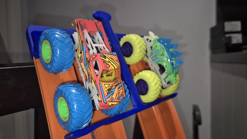 May include: Three toy monster trucks on a blue track. The trucks are a red and yellow monster truck, a green and white monster truck, and a blue and white monster truck.