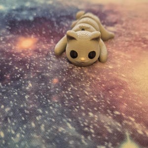 May include: A small, grey, 3D printed cat figurine with black eyes. The cat is curled up in a sleeping position. The figurine is on a galaxy-themed background.
