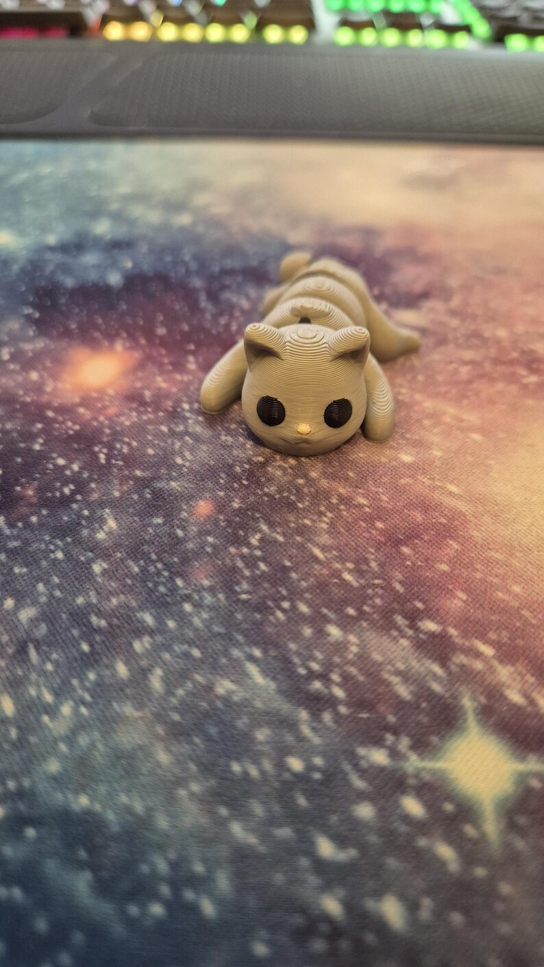 May include: A small, grey, 3D printed cat figurine with black eyes. The cat is curled up in a sleeping position. The figurine is on a galaxy-themed background.