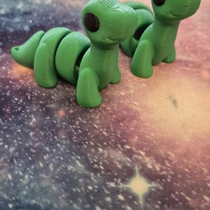May include: Two green, segmented, 3D printed toy dinosaurs with black eyes. The dinosaurs are posed in a playful, upright position.