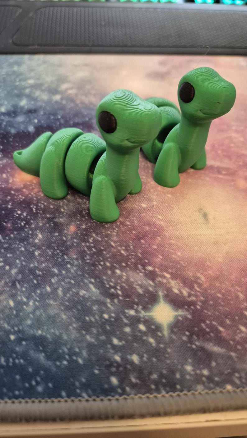 May include: Two green, segmented, 3D printed toy dinosaurs with black eyes. The dinosaurs are posed in a playful, upright position.