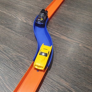 May include: A yellow toy car races down a blue track, followed by a black toy car on an orange track.