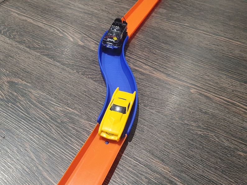 May include: A yellow toy car races down a blue track, followed by a black toy car on an orange track.