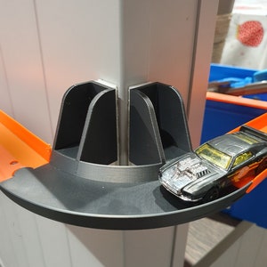 May include: A black toy car on a black and orange plastic track. The track is attached to a white wall with a black plastic connector.