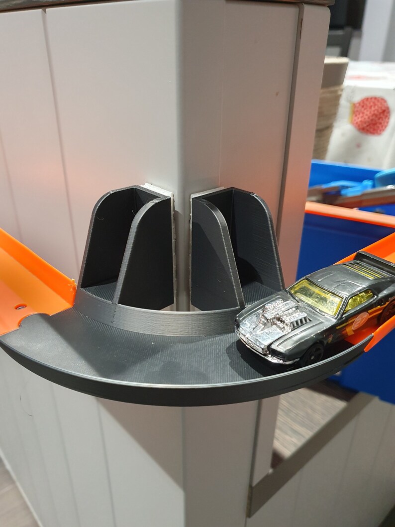 May include: A black toy car on a black and orange plastic track. The track is attached to a white wall with a black plastic connector.
