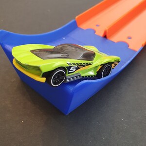 May include: A green toy race car with black wheels and a number 5 on the side is on a blue and orange plastic track.