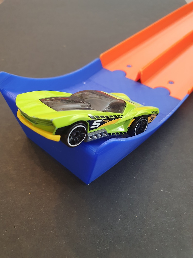 May include: A green toy race car with black wheels and a number 5 on the side is on a blue and orange plastic track.