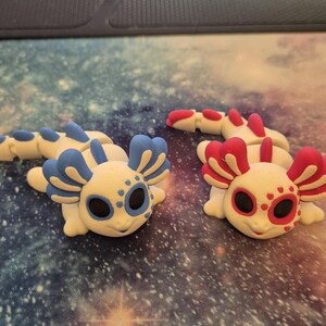 May include: Two small, white axolotl figurines with blue and red accents. The axolotls have black eyes and are posed on a galaxy-patterned surface.