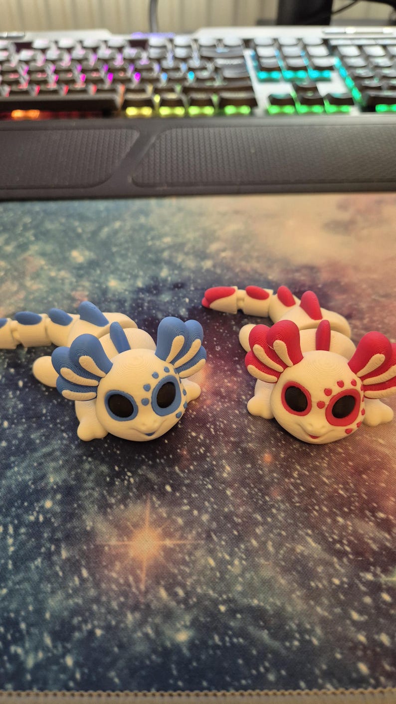 May include: Two small, white axolotl figurines with blue and red accents. The axolotls have black eyes and are posed on a galaxy-patterned surface.