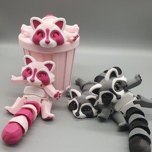 May include: Three 3D printed raccoon figurines. One pink raccoon is sitting in a pink trash can. Two gray raccoons are lying on their sides with their tails curled up.