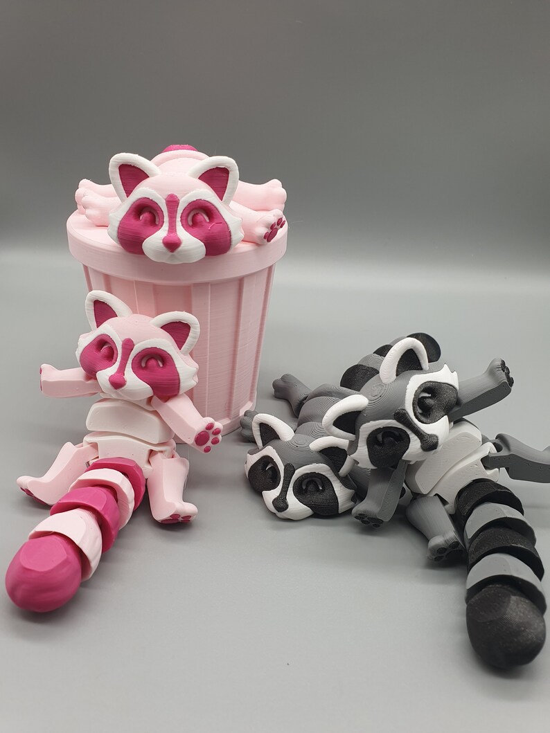 May include: Three 3D printed raccoon figurines. One pink raccoon is sitting in a pink trash can. Two gray raccoons are lying on their sides with their tails curled up.