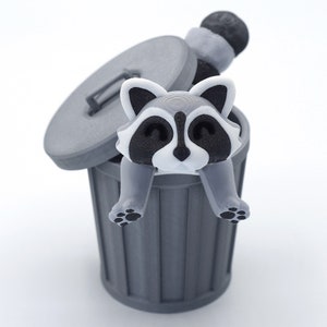 May include: A gray raccoon figurine with black eyes and a white nose is sitting inside a gray trash can with the lid open. The raccoon is holding onto the edge of the can with its paws.