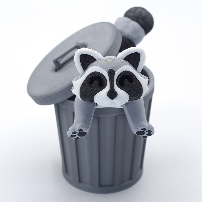 May include: A gray raccoon figurine with black eyes and a white nose is sitting inside a gray trash can with the lid open. The raccoon is holding onto the edge of the can with its paws.