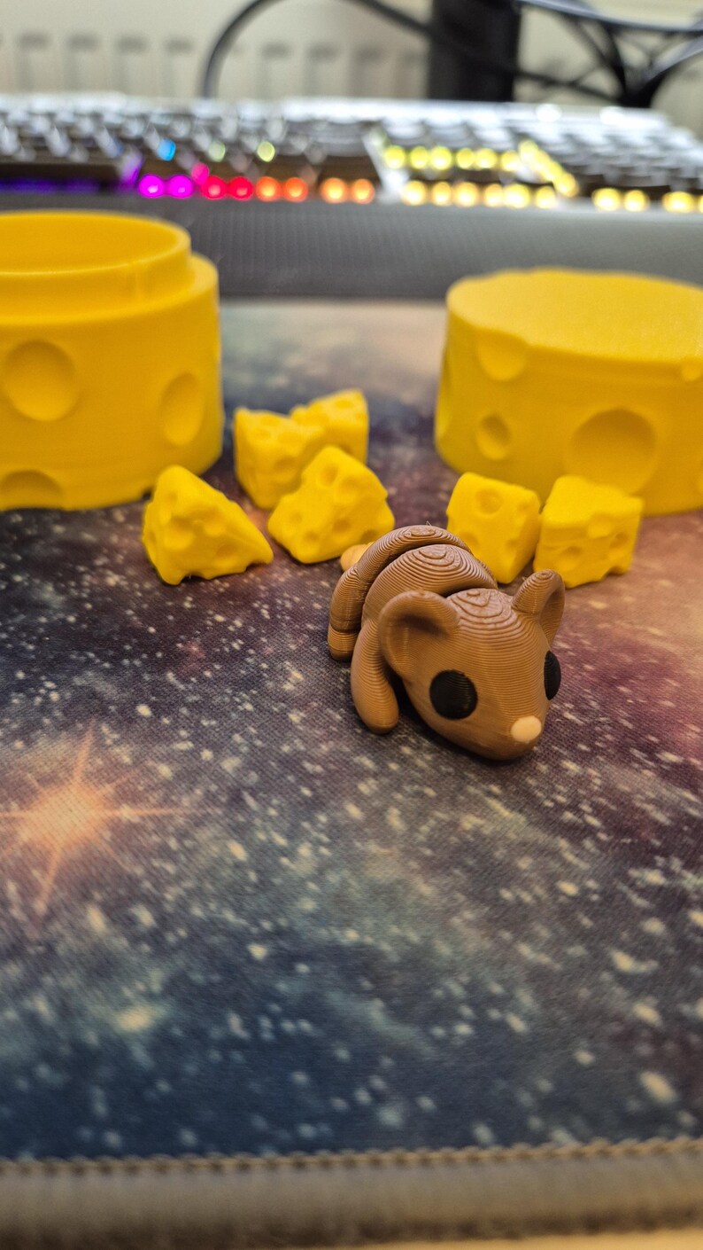 Puede incluir: Una figurita de rat&oacute;n impresa en 3D de color marr&oacute;n con ojos negros y nariz rosa, rodeada de bloques de queso amarillo.
