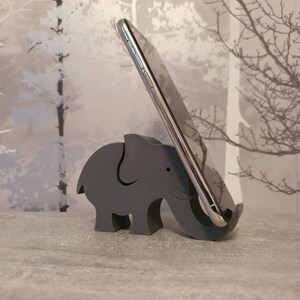 K&ouml;nnte beinhalten: Ein grauer Telefonst&auml;nder in Form eines Elefanten aus Holz. Der St&auml;nder h&auml;lt ein silbernes Smartphone. Der Elefant hat einen Ausschnitt, an dem das Telefon anliegt. Der Hintergrund hat ein Baumdesign.