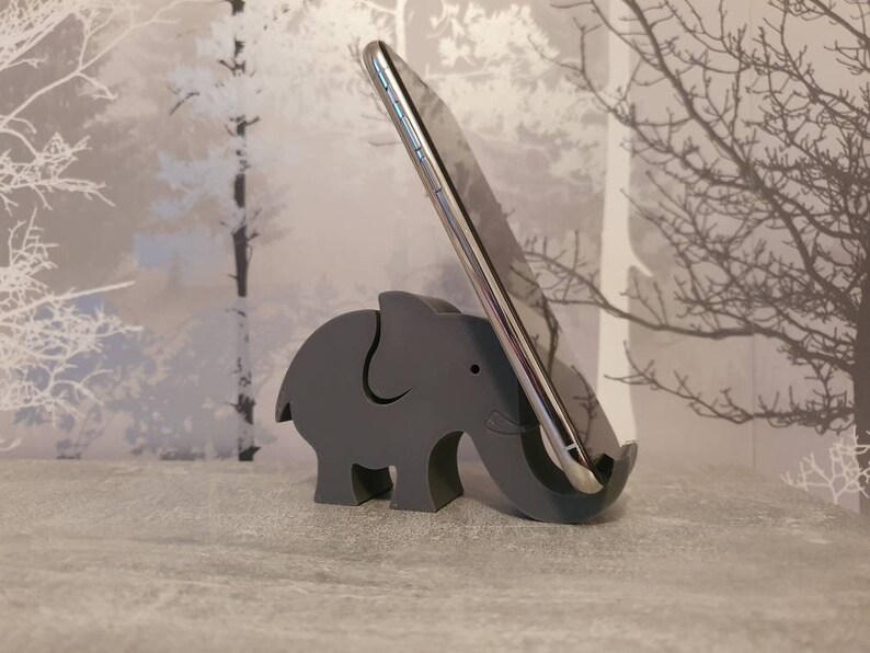 K&ouml;nnte beinhalten: Ein grauer Telefonst&auml;nder in Form eines Elefanten aus Holz. Der St&auml;nder h&auml;lt ein silbernes Smartphone. Der Elefant hat einen Ausschnitt, an dem das Telefon anliegt. Der Hintergrund hat ein Baumdesign.