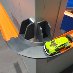 May include: A green toy car with black accents is driving on a gray and orange track. The track is part of a larger toy set, with a gray and white wall in the background.