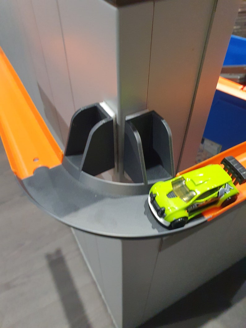 May include: A green toy car with black accents is driving on a gray and orange track. The track is part of a larger toy set, with a gray and white wall in the background.