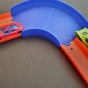 May include: A blue plastic track piece for a toy car race track. The track piece is curved and has a raised edge. Two toy cars are on the track, one red and one green.