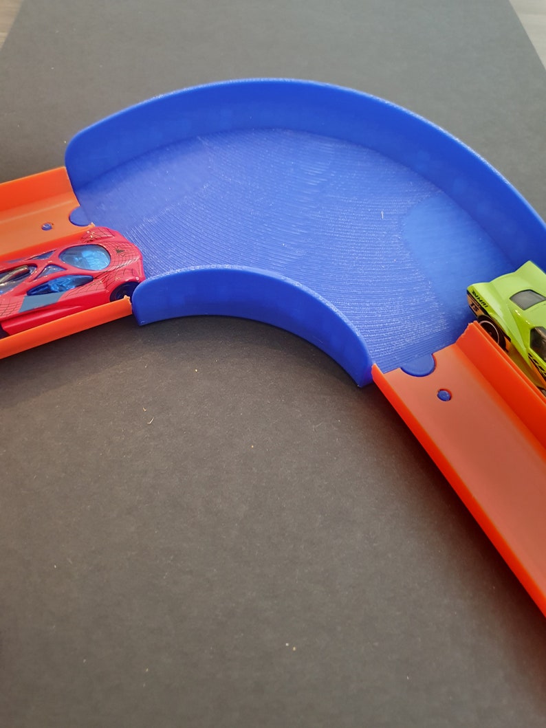 May include: A blue plastic track piece for a toy car race track. The track piece is curved and has a raised edge. Two toy cars are on the track, one red and one green.