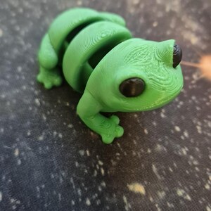 May include: A green 3D printed frog figurine with black eyes. The frog is sitting on a gray surface with white speckles.
