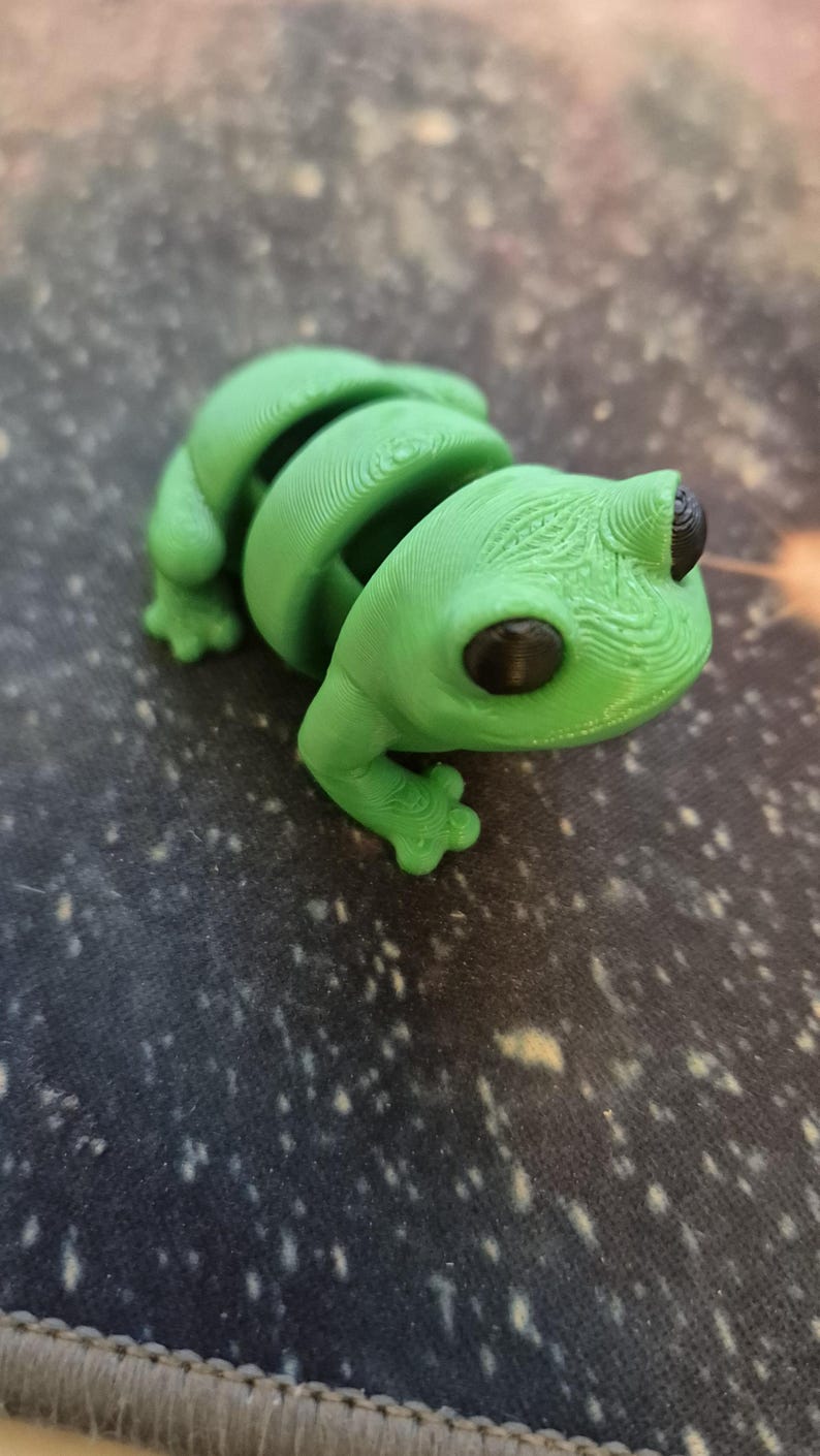May include: A green 3D printed frog figurine with black eyes. The frog is sitting on a gray surface with white speckles.