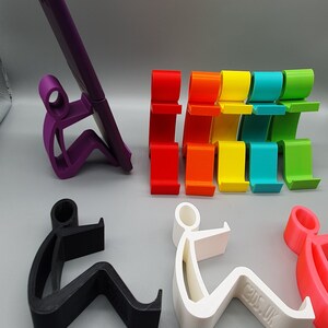 May include: A set of colorful phone stands in the shape of a person sitting down. The stands are made of plastic and come in a variety of colors, including red, orange, yellow, green, blue, and purple. There is also a black and white phone stand.