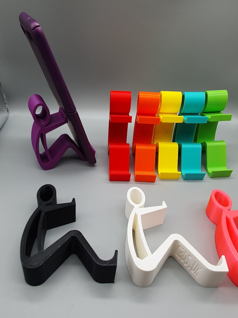May include: A set of colorful phone stands in the shape of a person sitting down. The stands are made of plastic and come in a variety of colors, including red, orange, yellow, green, blue, and purple. There is also a black and white phone stand.
