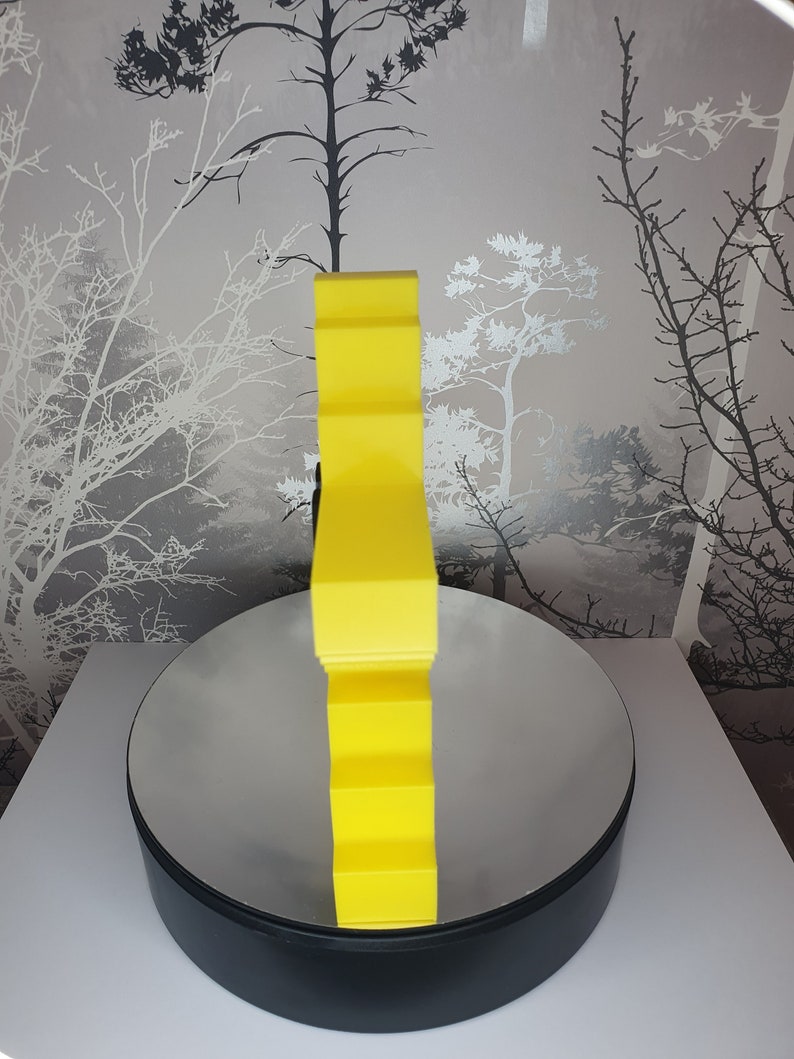 Puede incluir: Una escultura abstracta geom&eacute;trica amarilla con una forma ondulada y org&aacute;nica. La escultura est&aacute; sobre una base circular negra con una superficie reflectante.