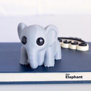 Op de afbeelding: Een grijze baby-olifantenfiguur met grote zwarte ogen staat op een blauw boek. De tekst "Baby Elephant" staat op het boek. Achter de olifant staat een zwart-wit object.
