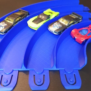 May include: Four toy cars on a blue plastic track. The cars are black, green, red, and gold. The track is curved and has four sections.