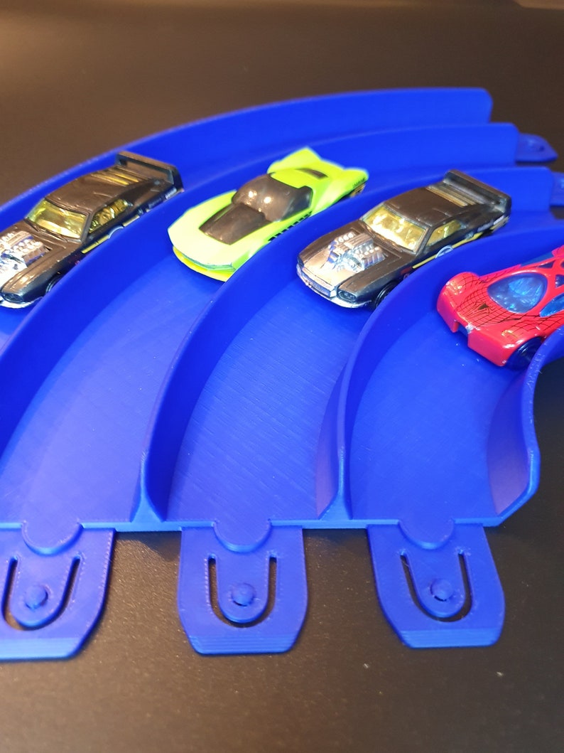 May include: Four toy cars on a blue plastic track. The cars are black, green, red, and gold. The track is curved and has four sections.